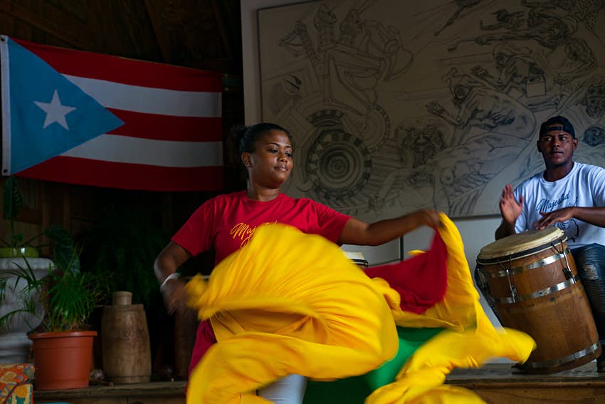 Una mujer gira su falda de colores brillantes hacia un hombre que toca un tambor en el fondo;  Puerto Rico Sustentable 